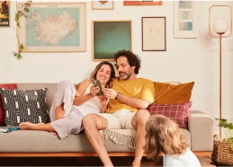 imagem de uma família composta por uma mulher, um homem e uma criança sentados em um sofá em uma sala de estar com diversos quadros na parede