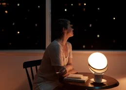 imagem de uma mulher sentada em uma cadeira diante de uma mesa com uma iluminária redonda em uma sala com meia luz em frente a uma janela durante a noite