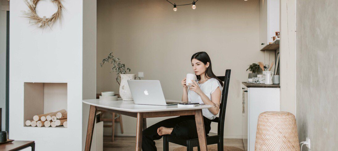 Foto que ilustra matéria sobre como investir em imóveis com uma mulher em casa, sentada em uma cadeira e tomando um café, assistindo um vídeo no computador.