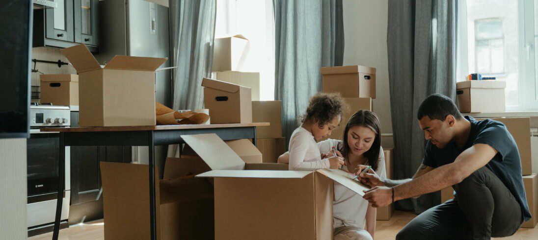 Foto que ilustra matéria sobre alta temporada de aluguel de imóveis, com um casal de um homem e uma mulher e uma criança entre várias caixas de mudança dentro de uma casa nova.