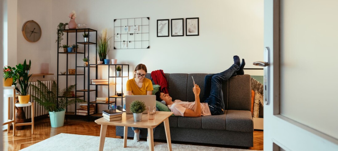 Foto que ilustra matéria sobre apartamento mobiliado para alugar mostra uma mulher e um homem em uma sala com quadros e plantas. A mulher mexe em um notebook, enquanto o homem está deitado no sofá.