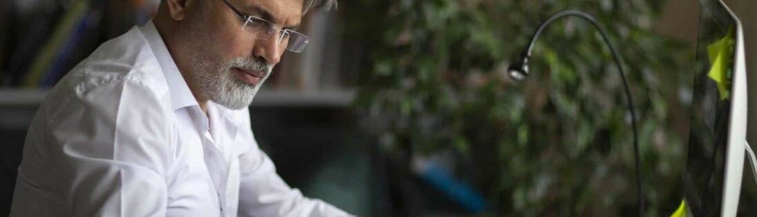 Homem de barba e camisa branca e usando óculos observa um computador ilustrando artigo sobre as novas funcionalidades do QuintoAndar para investimento em imóveis