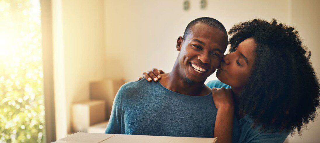 Foto que ilustra matéria sobre proposta de locação mostra um casal jovem que acaba de se mudar para uma casa nova. Na imagem, o homem sorridente ganha um beijo de sua parceira na bochecha.