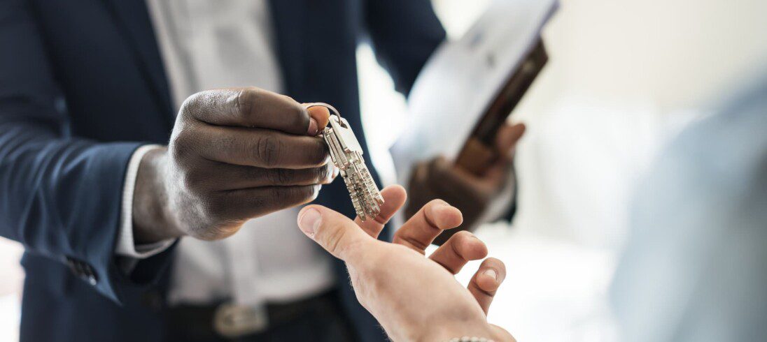 Em matérias sobre documentos para venda de imóvel, foto mostra detalhe da mão de um homem de terno entregando as chaves de um imóvel na mão de outra pessoa.