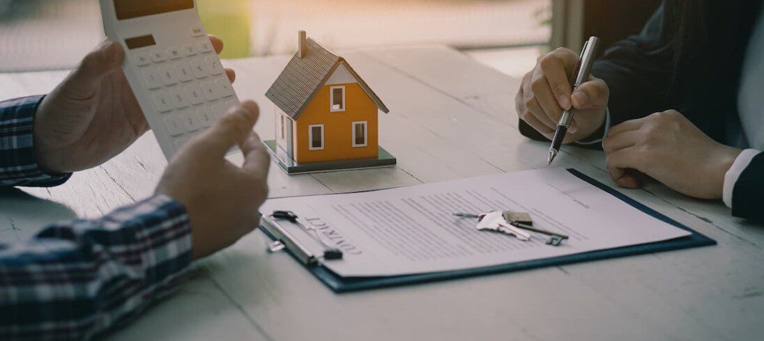 Pessoas analisam um contrato imobiliário com o auxílio de uma calculadora. Há, ao lado, a miniatura de uma casa.