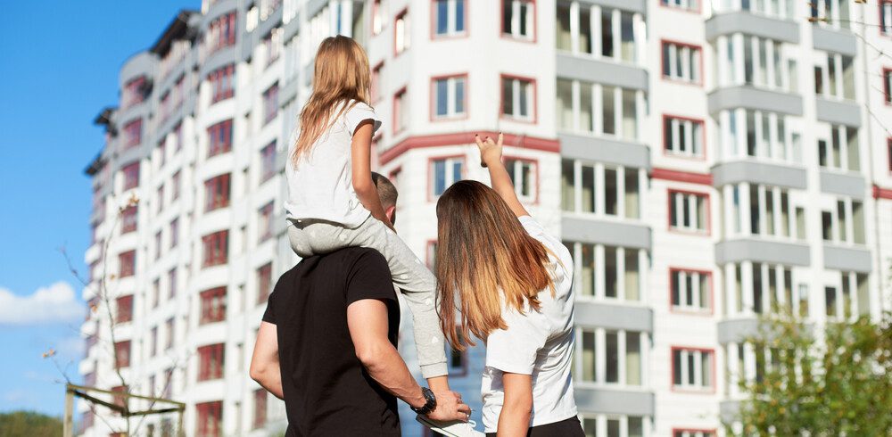 Em foto que ilustra matéria sobre como comprar um apartamento, um casal de costas aponta para um prédio ao fundo. A mulher é loira e o homem tem uma menina sentada em seus ombros.