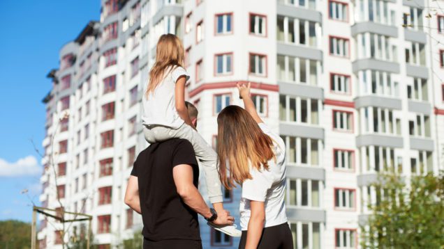 Em foto que ilustra matéria sobre como comprar um apartamento, um casal de costas aponta para um prédio ao fundo. A mulher é loira e o homem tem uma menina sentada em seus ombros.
