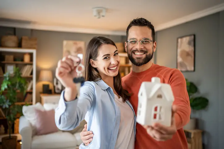 Imagem de um homem e uma mulher em uma casa segurando uma casa em miniautura e uma chave enquanto sorri para ilustrar matéria sobre como declarar imóvel financiado