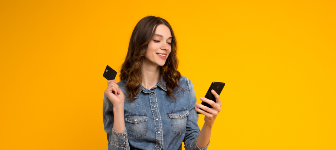 Foto que ilustra matéria que explica como pagar aluguel com cartão de crédito mostra uma mulher jovem olhando para o celular com um cartão de crédito na mão, com um fundo amarelo atrás dela.