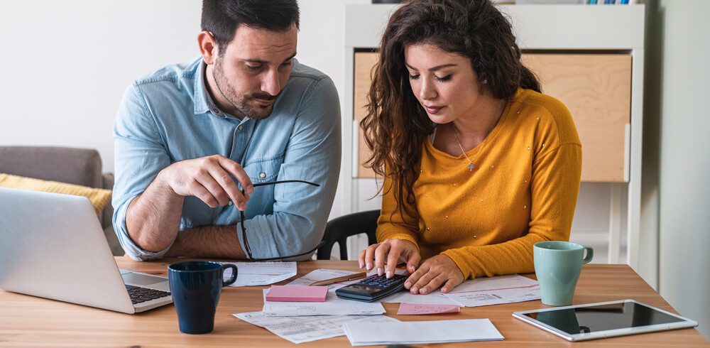 Foto que ilustra matéria sobre alugar ou financiar mostra um casal sentado a uma mesa, com um laptop e uma calculadora fazendo contas.