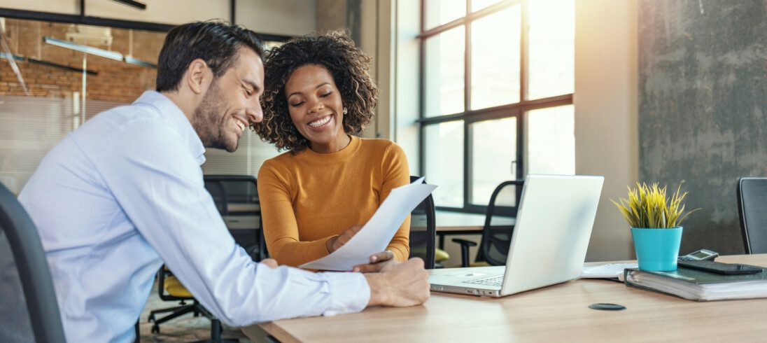 Em foto que ilustra matéria sobre avaliação de crédito, um homem e uma mulher sentados a mesa, com um laptop ao lado, sorriem enquanto olham para uma folha de papel