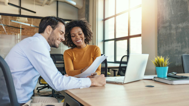 Em foto que ilustra matéria sobre avaliação de crédito, um homem e uma mulher sentados a mesa, com um laptop ao lado, sorriem enquanto olham para uma folha de papel