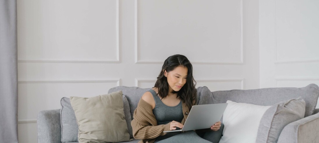 Foto que ilustra matéria sobre quando inquilino paga o primeiro aluguel, com uma mulher sentada no sofá pesquisando no computador.