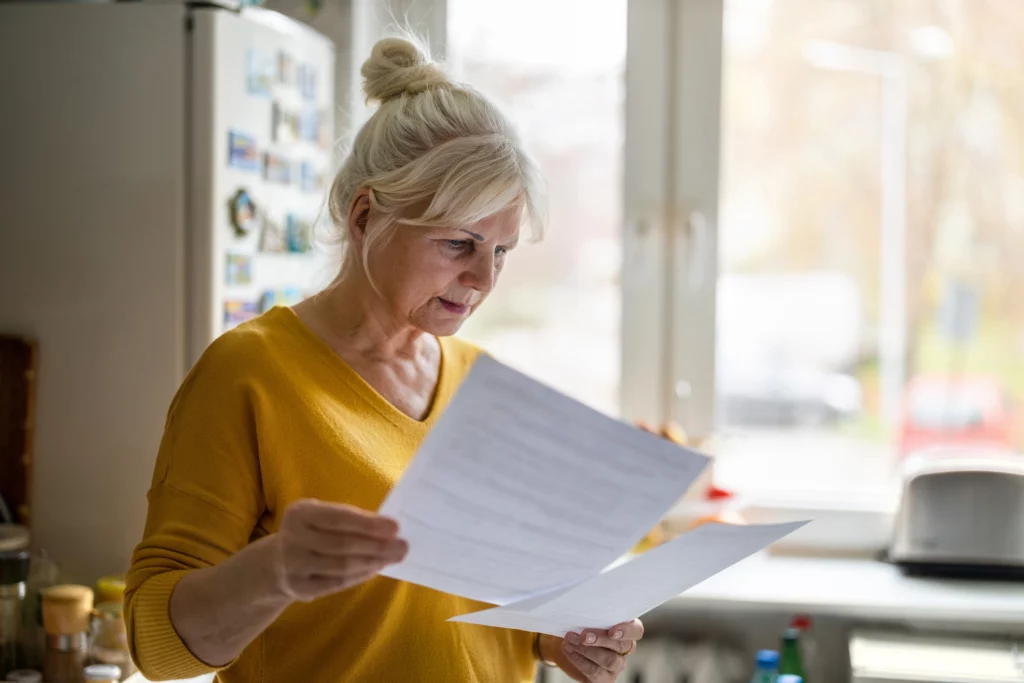 Imagem de uma mulher de mais idade e com o cabelo branco analisando alguns papéis na cozinha de casa para ilustrar matéria sobre como funciona o seguro fiança para alugar um imóvel
