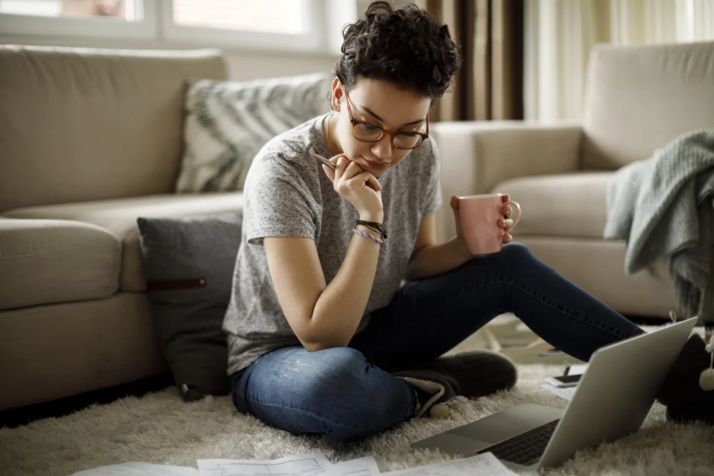 Imagem de uma mulher com o cabelo curto sentada em cima do tapete da sala em frente a um sofá olhando para a tela do computador que está no chão para ilustrar matéria sobre seguro fiança