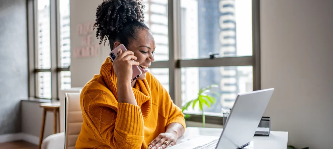 Imagem de uma mulher negra vestindo casaco laranja sentada na mesa da sala de casa olhando para a tela do computador sorrindo e atendimento ao telefone celular para ilustrar matéria sobre o seguro fiança locatícia