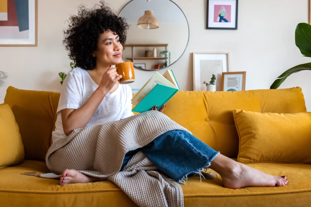 Imagem de uma mulher sentada em um sofá amarelo na sala de estar de uma casa com um livro azul e uma caneca amarela nas mãos para ilustrar matérias sobre o seguro fiança para aluguel