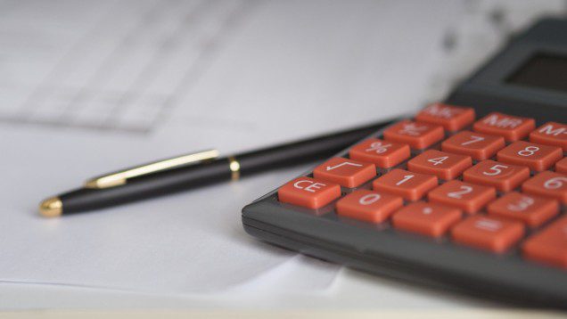 Foto que ilustra matéria sobre juros imobiliários mostra uma calculadora com botões vermelhos em destaque, com uma caneta preta ao lado, ambas em cima de alguns papéis