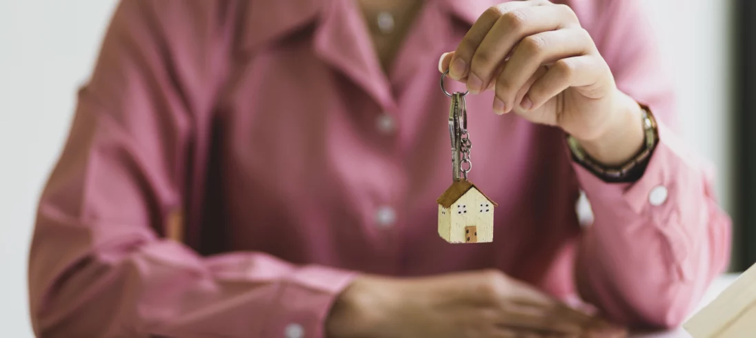 Imagem do corpo de uma mulher de camiseta rosa como foco nas mãos, onde ela está segurando uma chave com um chaveiro em formato de casa para ilustrar matéria sobre casa ou apartamento