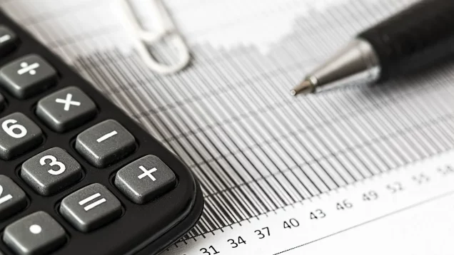 Foto que ilustra matéria sobre aplicar em investimentos ou imóveis mostra parte de uma calculadora e uma caneta em primeiro plano, com uma folha de papel de fundo.