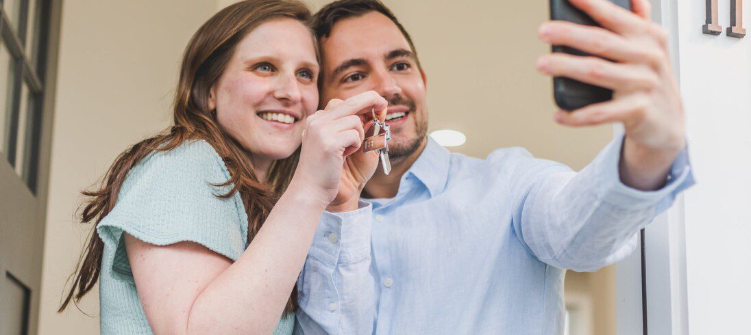 Foto que ilustra matéria sobre como montar uma carteira de investimentos para comprar um imóvel mostra um jovem casal tirando uma foto com o celular mostrando as chaves da casa nova