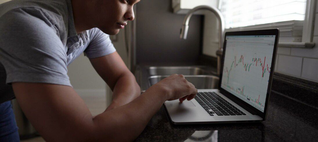 Foto que ilustra matéria sobre renda passiva mostra um homem jovem de pé e curvado sobre uma bancada de cozinha, que tem uma torneira ao fundo. Ele olha para um computador cheio de gráficos, que representam investimentos.