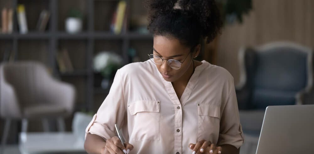 Foto que ilustra matéria sobre juros de mora mostra uma mulher negra, sentada a uma mesa, com um notebook aberto de lado, fazendo contas em uma calculadora e escrevendo em um papel.