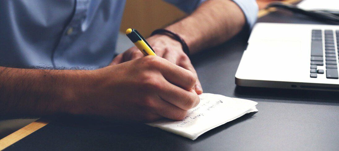 Foto de um homem sentado, de frente para um notebook e fazendo anotações em uma folha de papel com uma caneta.