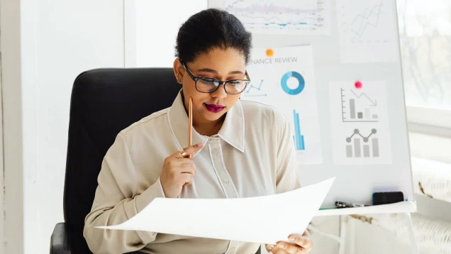 Foto que ilustra matéria sobre mercado de imóveis usados aquecido mostra uma mulher de óculos olhando para uma folha de papel, com uma caneta na mão, demonstrando que está pensando, com gráficos ao fundo em uma lousa.