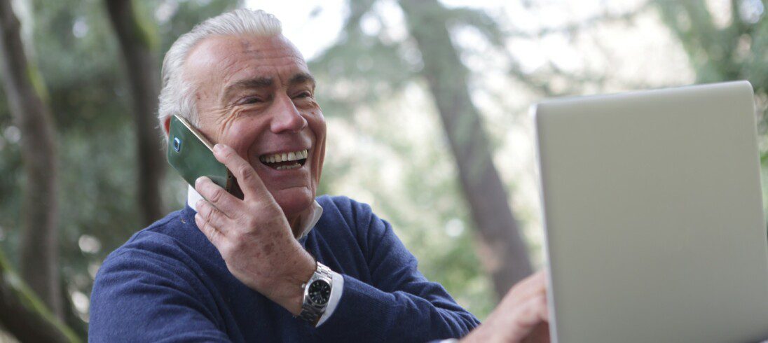 Foto de um homem sentado, de frente para um notebook. Ele está sorrindo e usando o celular.