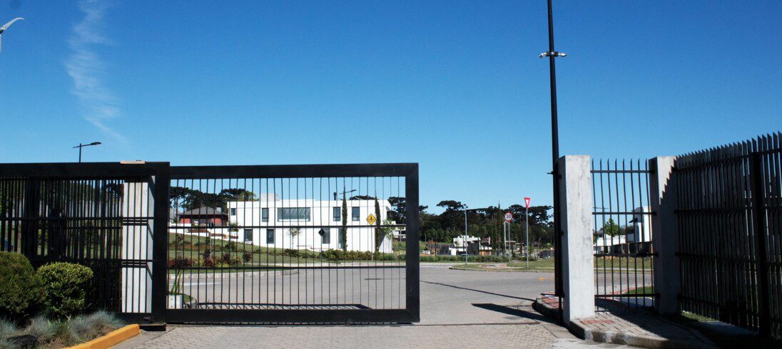 Foto que ilustra matéria sobre condomínios fechados mostra um portão de entrada para carros de um condomínio. Ao fundo, aparece uma casa branca