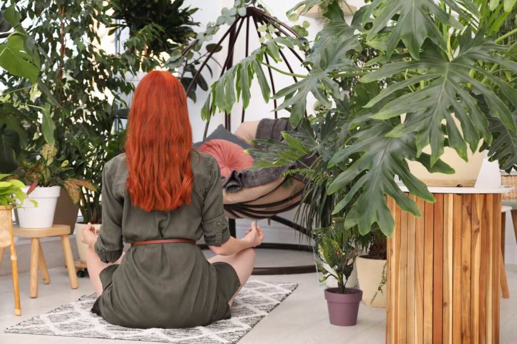 Mulher meditando no chão da varanda. A foto mostra um cenário com muitas plantas ao redor da mulher, que está de costas.