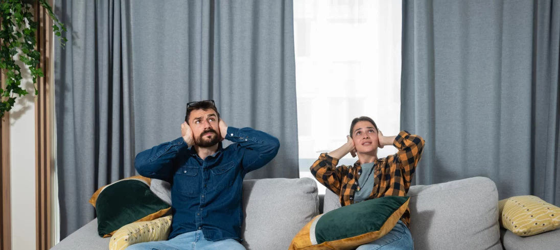 Imagem de um jovem casal, composto por um homem e uma mulher, sentado no sofá de casa olhando para cima com expressão de incômodo e com as mãos nos ouvidos para ilustrar matéria sobre perturbação do sossego