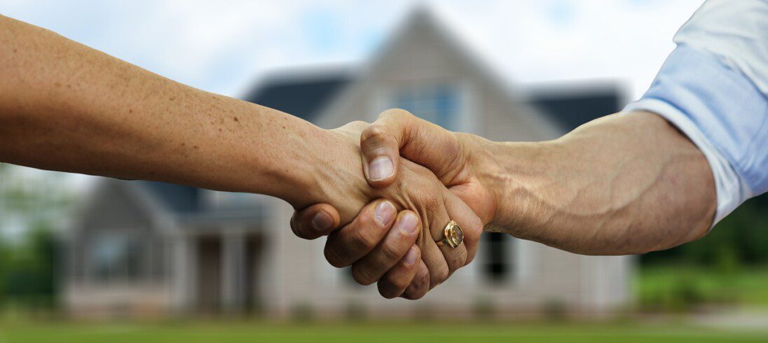 Foto que ilustra matéria sobre como escolher uma imobiliária para alugar e vender imóveis mostra duas pessoas apertando as mãos em frente a uma casa