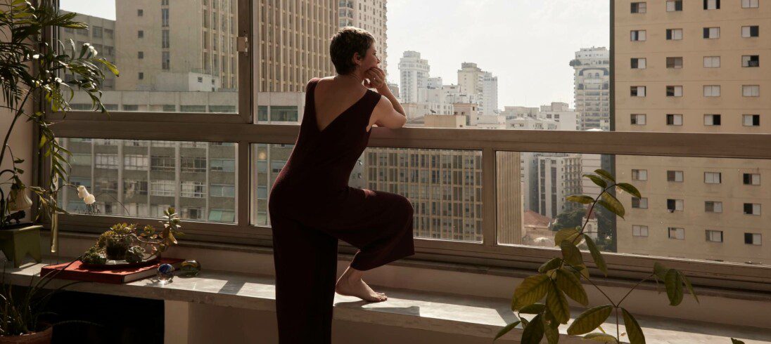 Foto que ilustra matéria sobre o mercado residencial na América Latina mostra uma mulher de costas, apoiada no peitoril de uma janela de um apartamento em um prédio alto olhando para outros prédios altos a sua frente.