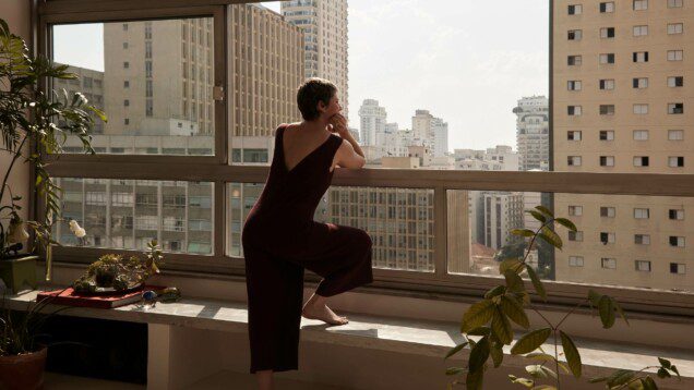 Foto que ilustra matéria sobre o mercado residencial na América Latina mostra uma mulher de costas, apoiada no peitoril de uma janela de um apartamento em um prédio alto olhando para outros prédios altos a sua frente.