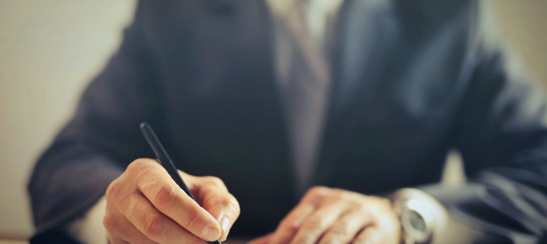 Homem com traje de executivo assina um papel com uma caneta preta. Imagem disponível em Pexels.