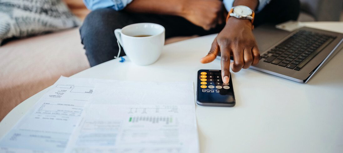 Foto que ilustra matéria sobre imóveis que geram renda mostra um homem fazendo contas em uma calculadora de celular que está em cima de uma mesinha com um computador ao lado e papéis