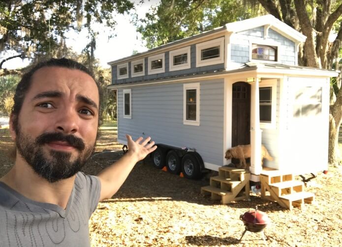Foto que ilustra matéria sobre tiny house mostra um homem - Robson, o entrevistado da matéria - fazendo uma selfie apontando para uma pequena casa sobre rodas atrás dele.