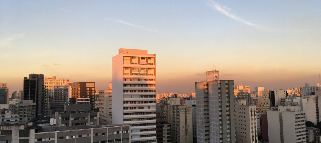 A imagem mostra a vista com prédios e casas no fim de tarde com o pôr do sol.