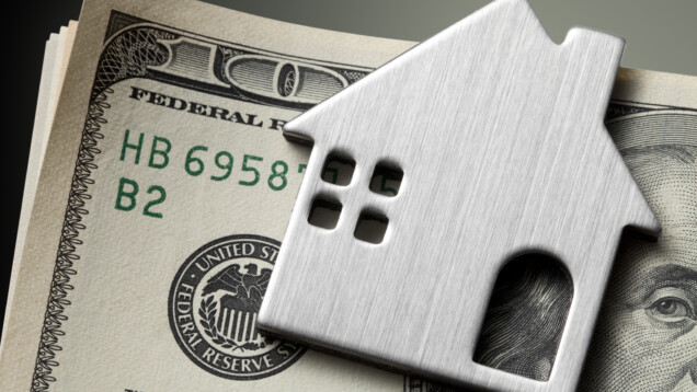 A imagem mostra uma nota de dólar com metal no formato de uma casa em cima.