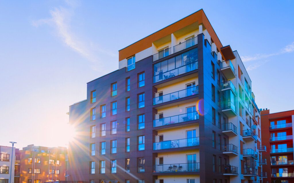 A imagem mostra a arquitetura de uma fachada residencial de um prédio com apartamentos. Os apartamentos possuem varandas ao ar livre e recebem o reflexo da luz solar.
