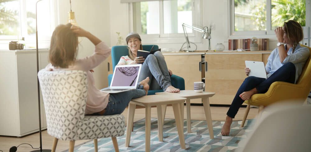 Foto que ilustra matéria sobre coliving mostra um grupo de três jovens sentadas em uma sala de estar. Elas conversam e riem. Duas delas têm notebooks em seus colos, enquanto a terceira tem um livro aberto nas mãos