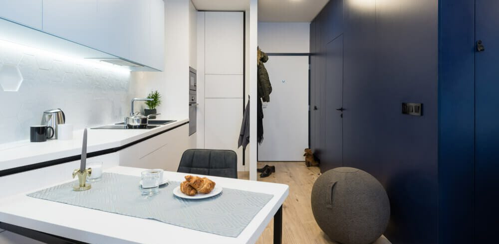Foto mostra o interior de um moderno microapartamento, com um espaço com bancada e armários de cozinha brancos, integrados a uma pequena mesa. Do lado oposto, uma parede azul com uma porta. E no meio, ao fundo, a porta de entrada do imóvel.