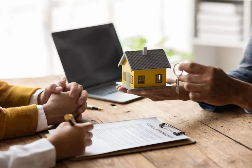 Imagem de três pessoas sentadas em uma mesa onde uma está mostrando uma casa em miniatura e as chaves de uma casa para as outras para ilustrar matéria sobre o impacto da TR no financiamento imobiliário