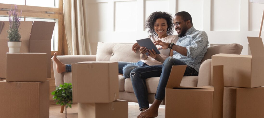 Foto de um homem e uma mulher negros sentados em um sofá, rodeados de caixas de mudança. Eles procuram por um novo lar em um tablet.