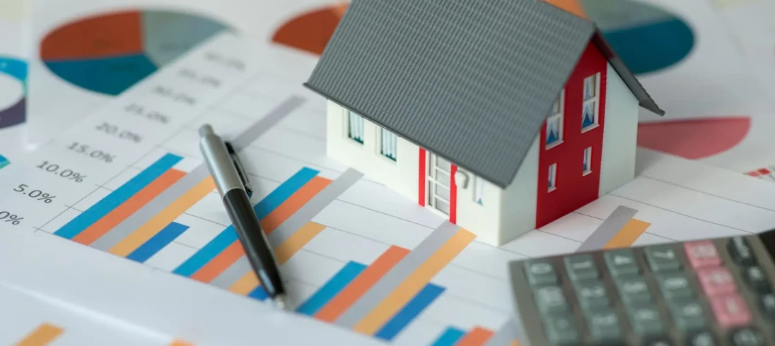 Imagem da miniatura de uma casa em cima de alguns papéis com gráficos e ao lado de uma calculadora para ilustrar matéria sobre taxa referencial