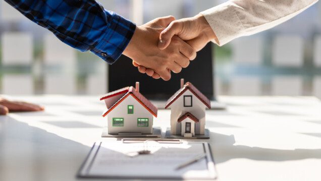 Foto que ilustra matéria sobre rentabilidade de imóveis mostra duas pessoas dando as mãos com uma caderneta e mini casinhas que estão na mesa.