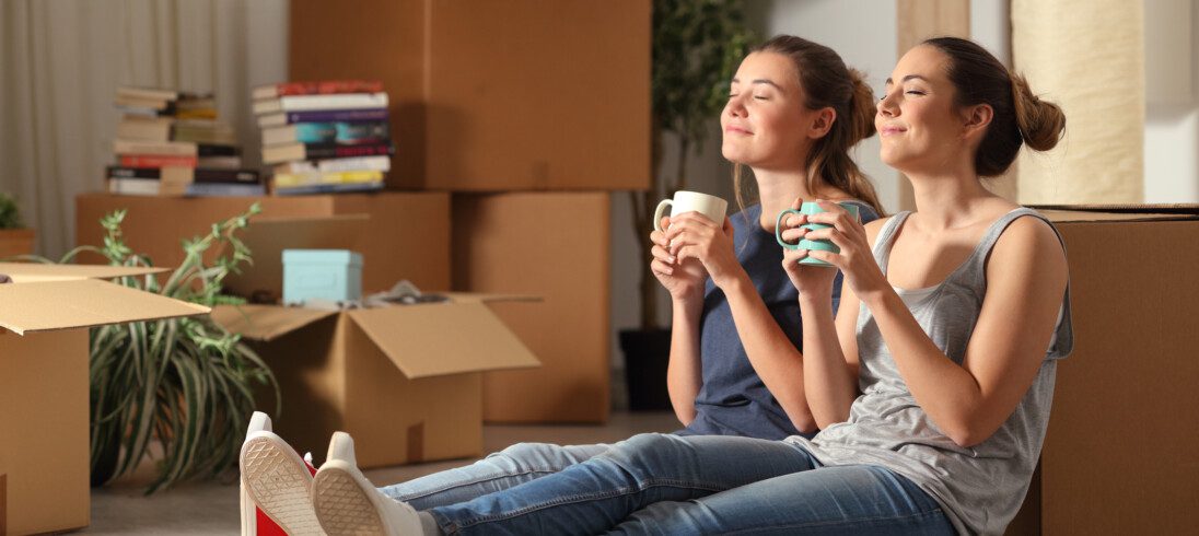 Foto que ilustra matéria sobre dividir aluguel mostra duas garotas sentadas lado a lado no meio de caixas de mudança segurando uma xícara nas mãos