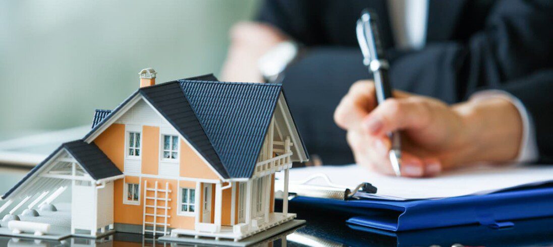 Uma pessoa assina um contrato de financiamento imobiliário. Em cima da mesa, ao lado do contrato, há uma miniatura de casa.